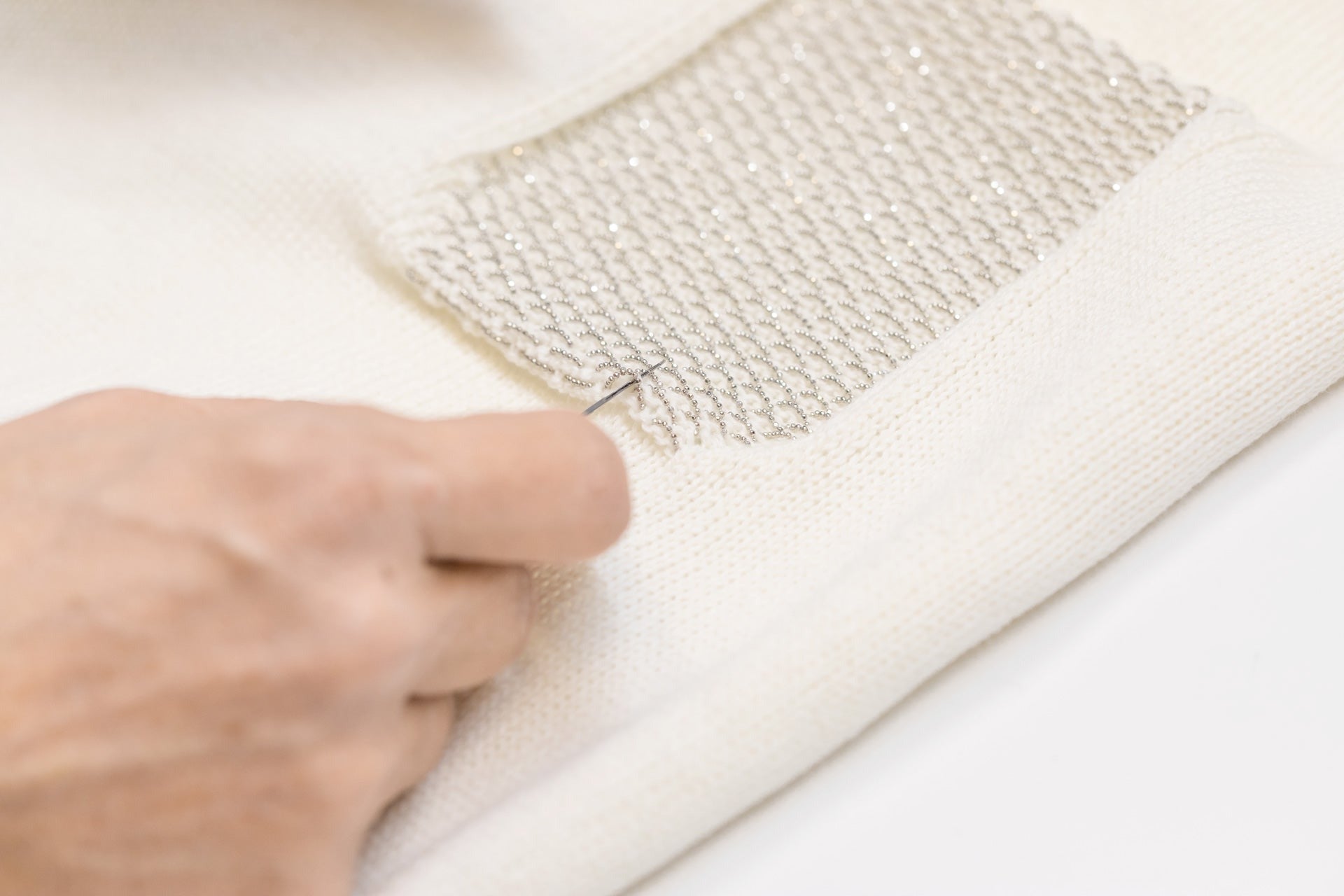 Handwerk van Peserico: een zakje wordt met een naald op een gebreide pullover manueel afgewerkt.