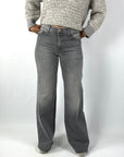 Een grijze damesjeans met middelhoge taille en wijde, recht vallende pijpen. De broek heeft voor- en achterzakken met decoratief stiksel en een witte zilveren knoopsluiting.